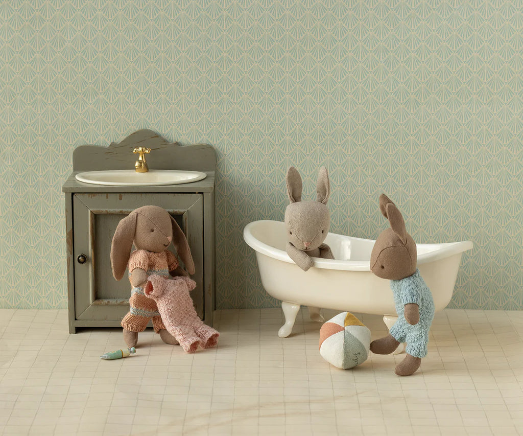 Three micro bunny toys play in a miniature bathroom scene with a bathtub, sink, and pastel ball. One bunny is relaxing in the tub, one stands nearby, and one wears the Maileg Extra Clothing: Knitted Suit, Micro - Rose for Micro Bunny.