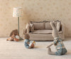 Three Maileg Micro Bunnies With Ball (Brown, Dusty Rose, Magnetic Hands) sit on a light carpet before a beige sofa; two hold soft balls. A floral lampshade and floral-patterned wallpaper complete the cozy scene.