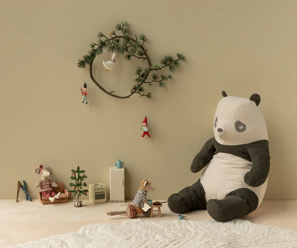 A cozy room features the Maileg Metal Ornament - Pixy, Candle Light hanging from a pine branch on a beige wall, alongside a large stuffed panda, small animal dolls, a miniature kitchen set, and a soft white rug.