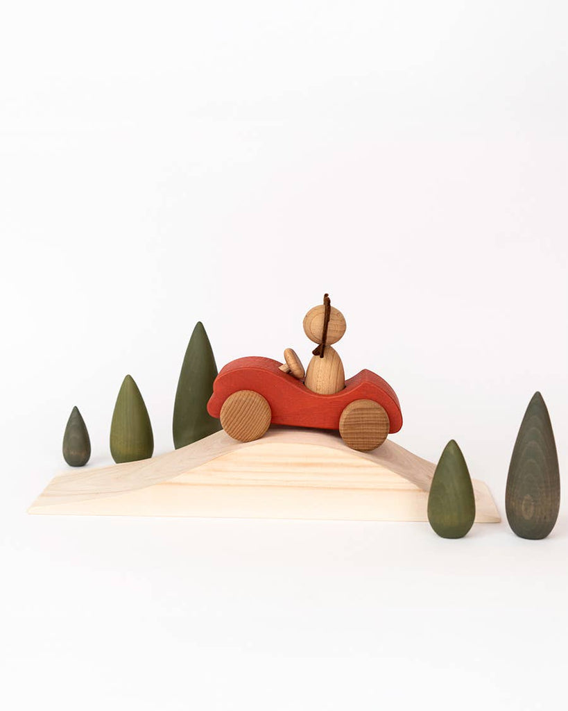 A wooden figure drives a red car up a ramp, surrounded by minimalist green trees on the Sabo Concept First Journey play set—perfect for inspiring imaginative, open-ended play against a white background.