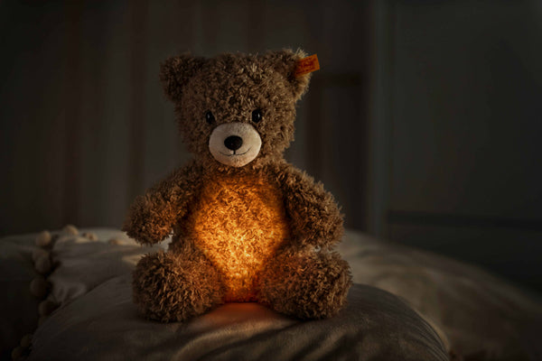 In a dim room, the Light at Night Caspar Teddy Bear Stuffed Plush, 11 inches, glows warmly like a cozy night light. Its fluffy brown fur shines softly, and a red tag hangs from its left ear. A USB charger stands by to power its gentle glow.