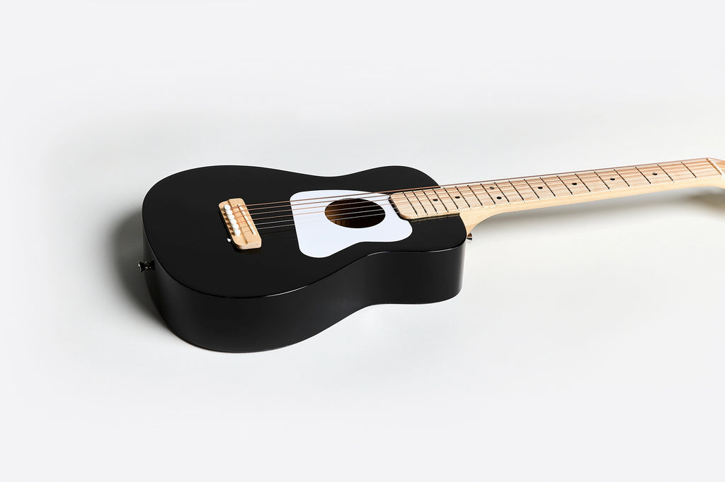 A Loog Pro Vi Acoustic Guitar Ages 9+ (ships in approximately one week) with a light wood neck and fretboard lies on a white surface. The guitar features a white pickguard and gold-colored bridge pins, positioned diagonally from the bottom left to the top right of the image.