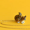 A small porcelain figurine of a brown and white cat, hand painted, with a 24k gold plated Squirrel Necklace attached, sitting on a plain yellow background with a golden chain loosely laid next to