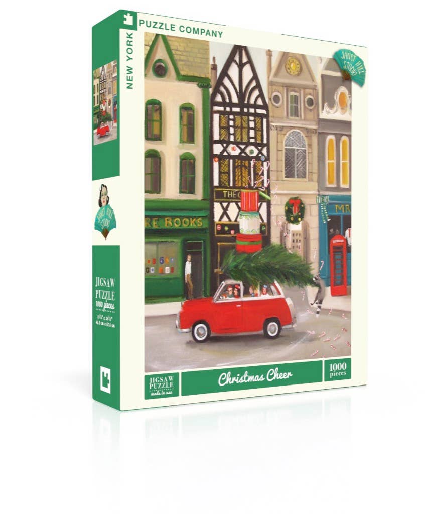 A 1000-piece Christmas Cheer - 1000 Piece Jigsaw Puzzle box featuring a Janet Hill illustration of a red car carrying a Christmas tree in front of storefronts decorated for the holidays.
