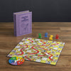 The Chutes and Ladders Vintage Bookshelf Edition features a purple book-shaped box, a vibrant board, spinner, and four colored pieces on a wooden table—perfect for enjoying the classic game’s playful twists and turns.