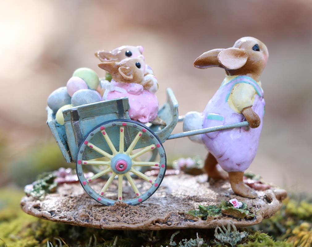 The Wee Forest Folk Tiny Collectible - Easter's on its Way (Not a Toy) is a hand-sculpted miniature featuring a bunny in pastels pushing a blue wheelbarrow with colorful eggs and two small bunnies, set on a floral base.