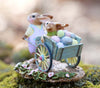 The Wee Forest Folk Tiny Collectible - Easter's on its Way (Not a Toy) features a hand-sculpted rabbit pushing a blue wheelbarrow with pastel eggs and a baby bunny on a mossy, flowered base—ideal for Wee Forest Folk fans.