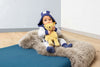 A baby dressed in a cozy animal-themed hoodie and socks sits on a fluffy rug, happily holding a yellow "My First Steiff" teddy bear. The background is minimal with a plain wall and Steiff, My First Steiff Teddy Bear, Beige, 10".