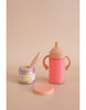 The My Tiny Birthday Set - Tiny Harlow includes a whimsical small jar called "tiny tummies" and a pink two-handled sippy cup, perfect for realistic play and delightful imaginary tea parties with birthday cake jars and strawberry milk, all set against a beige backdrop.