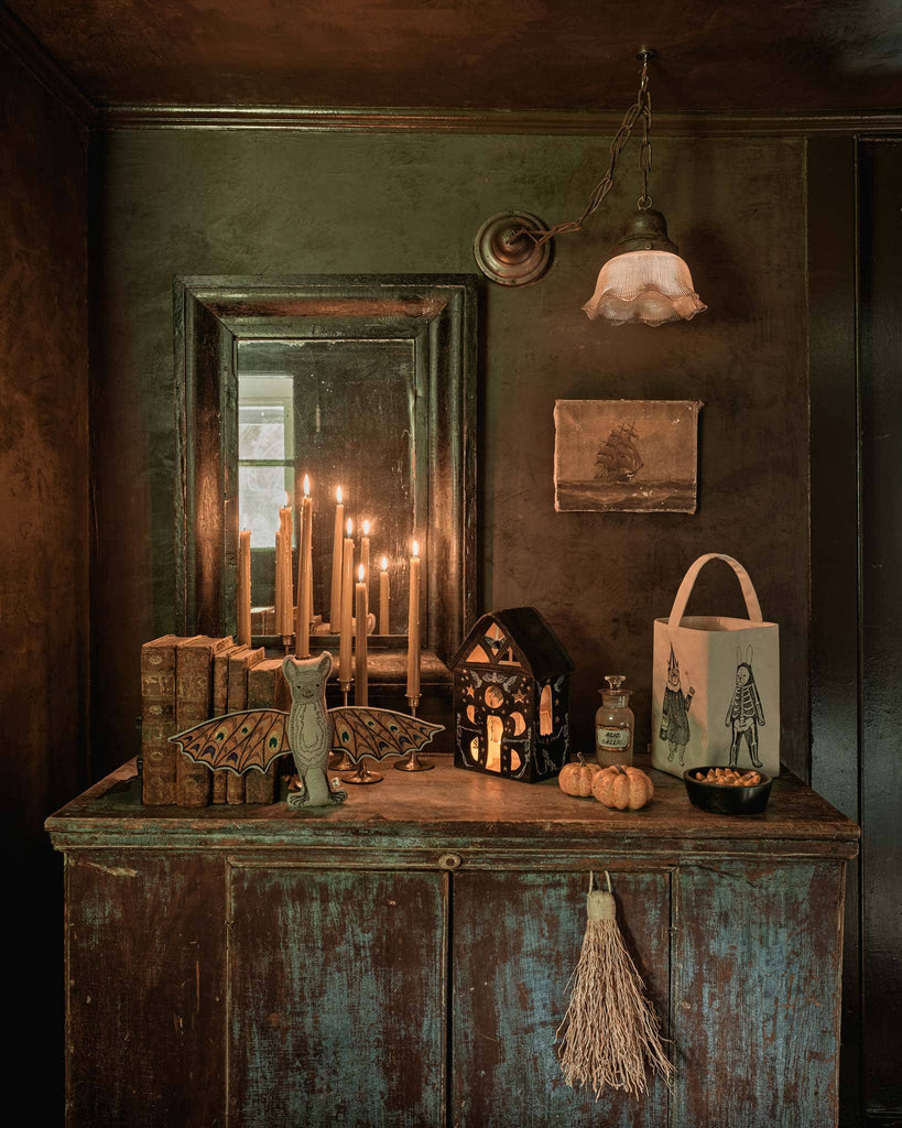 A rustic wooden cabinet showcases vintage books, lit candles, a haunted house lantern, a small pumpkin, broom, Halloween bag, dish, and the Coral & Tusk Bat Surprise Doll (ships in one week), creating a moody Halloween scene.