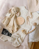 A flat lay of baby items features the Cuddle + Kind Organic Cotton Baby Bunny Lovey (Oatmeal), cream baby clothes, a wooden "hello world" sign, baby socks, and an ultrasound photo on a soft fabric background.