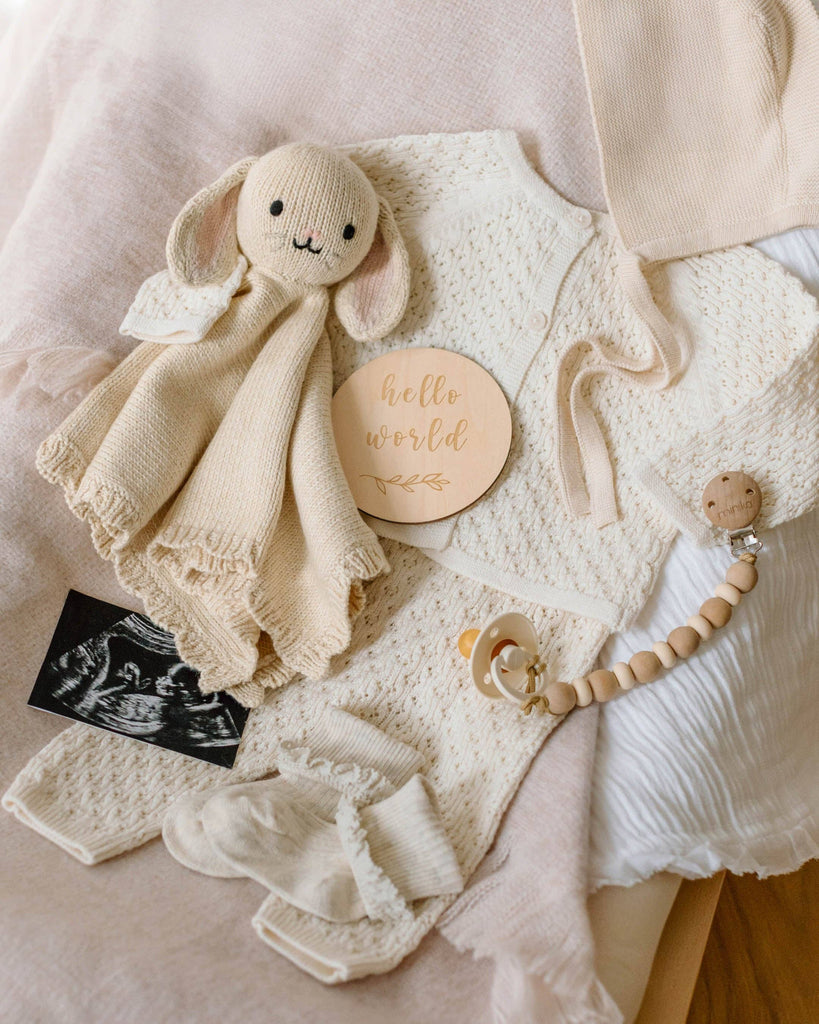 A flat lay of baby items features the Cuddle + Kind Organic Cotton Baby Bunny Lovey (Oatmeal), cream baby clothes, a wooden "hello world" sign, baby socks, and an ultrasound photo on a soft fabric background.