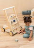 A toddler in blue overalls sits on a wooden floor next to the Wooden Activity Walker, which features colorful blocks and interactive elements. The child looks down, touching the floor and is surrounded by engaging sensory toys that encourage exploration and play.