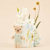 Cuddle + Kind Tiny Charlie the Honey Bear, a hand-knit doll in light blue overalls, stands in front of a floral-themed box with white and yellow flowers and light blue hydrangeas against a beige background.