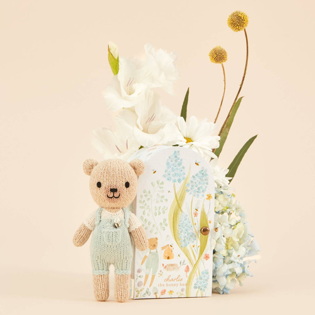 Cuddle + Kind Tiny Charlie the Honey Bear, a hand-knit doll in light blue overalls, stands in front of a floral-themed box with white and yellow flowers and light blue hydrangeas against a beige background.