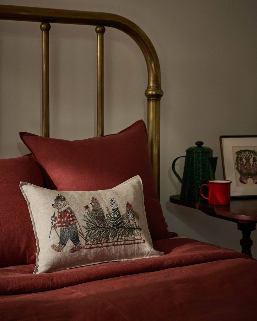 A Coral & Tusk Christmas Tree Sled Pillow, a rectangular holiday decorative pillow featuring an embroidered scene of animals pulling a Christmas tree on a wooden sled, resting on a bed with dark red bedding.