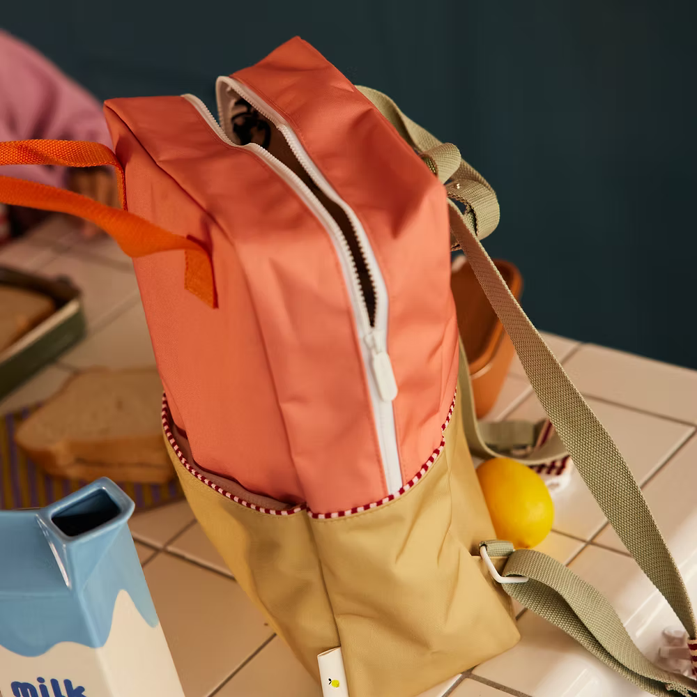 The Sticky Lemon Backpack Large | Color Blocking in Coral Pink + Reed Green, featuring orange handles and a green strap, sits on a tiled table by a lemon, milk carton, and bread slices. Its main pocket is open to show the waterproof lining and white zipper.