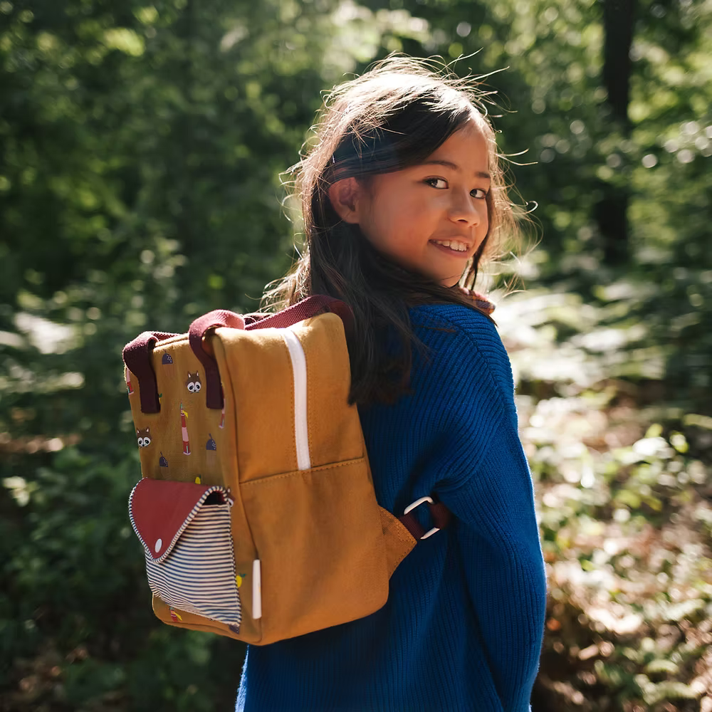 A young girl with long dark hair, wearing a blue sweater and carrying the Sticky Lemon Backpack Small Special Edition | Adventure Collection | Khaki Green by its shoulder handle, smiles on a sunlit path in a green forest.