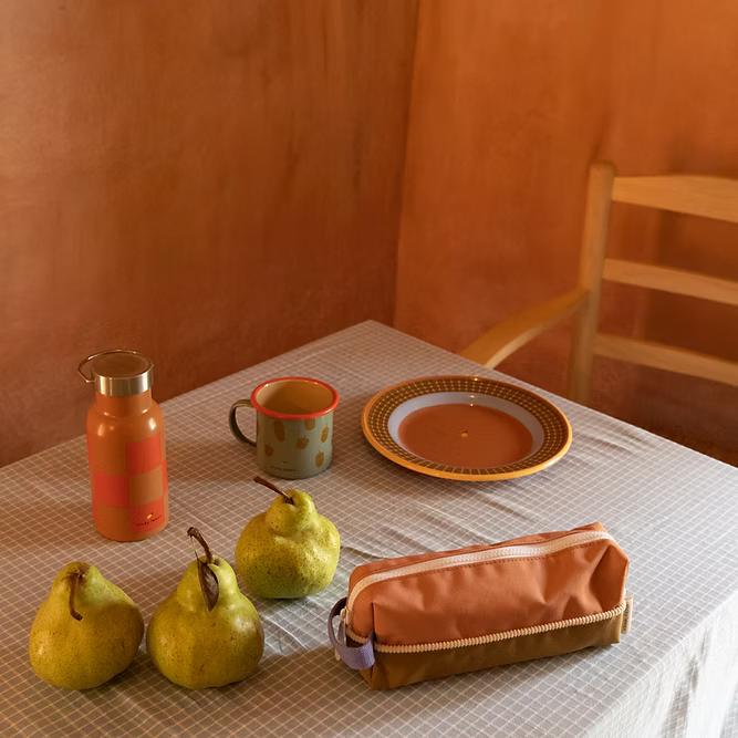 A table with a checkered cloth holds three pears, a pencil case, a Sticky Lemon Water Bottle | Farmhouse | Ladybird Red + Harvest Moon, a mug, and an empty plate. A wooden chair sits next to the table against a warm-toned wall.