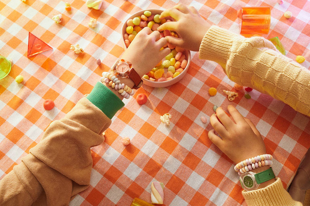 Two people with beaded bracelets reach for colorful snacks on an orange checkered tablecloth, both wearing a Mini Kyomo Watch - Cinnamon Roll. Scattered snacks and geometric shapes decorate the table.