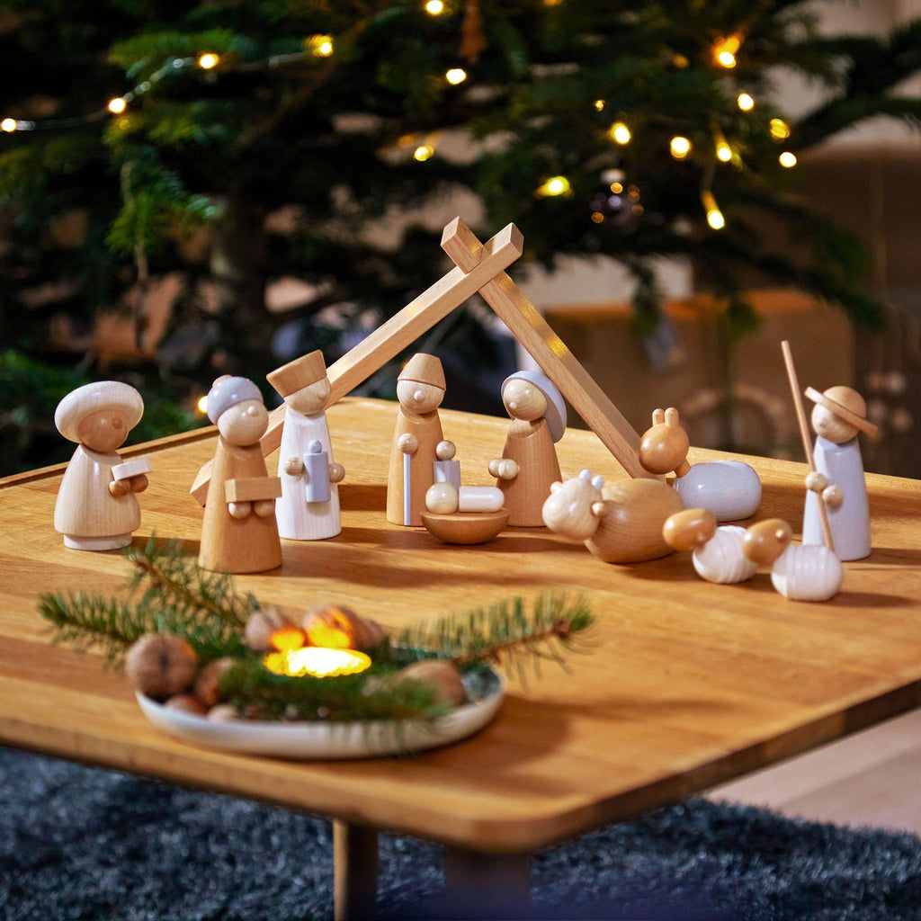 The Wooden Nativity Scene, crafted in Germany, is displayed on a table with Mary, Joseph, baby Jesus, animals, and more. A glowing Christmas tree stands behind it while a tray with candles and greenery adorns the foreground.