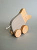 The Pull and Push Toy Little Wild Boar is a charming handcrafted toy featuring a wooden pig on wheels with a painted striped body, cheerful smile, and short string tail. Made from sustainable materials, it stands out beautifully against the plain gray background.