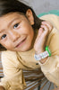 A young child with long hair, in a beige sweater and green Mini Kyomo Watch x Taxi Brousse - Color Notebook, smiles while leaning on a vintage wooden chair.