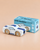 A white wooden toy car with blue stripes, inspired by iconic muscle car designs, is displayed next to its blue packaging box labeled “No. 17 GT95 Maverick | Made in England” on a beige surface.