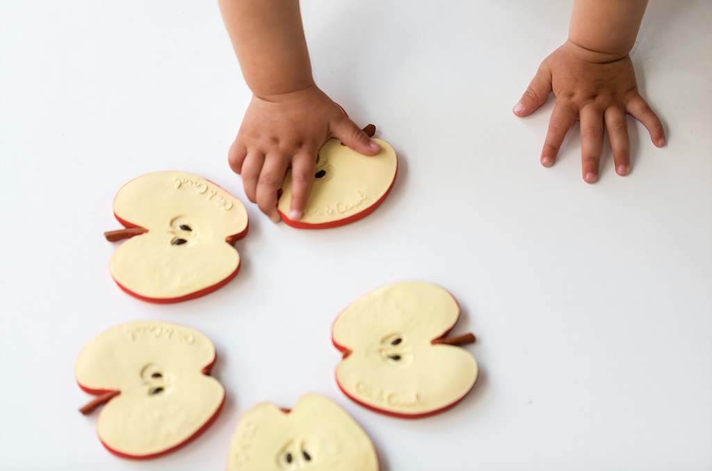 A child’s hands reach for four Pepita the Apple Teething Toys—apple slice-shaped teethers made from natural rubber—safe and designed for sensory play, not real fruit.