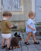Two toddlers walk on a cobblestone street, each pulling a charming Pull-Along Raoul on wheels. One wears a brown sweater and white shorts, the other a light blue outfit. A worn, shuttered window and a gray door are in the background.