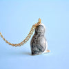 A Tiny Penguin Necklace, photographed against a blue background. The handmade necklace features the penguin facing away, showcasing its gray and white textured back.