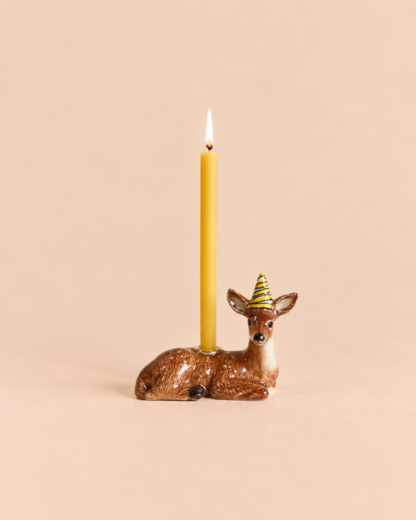 The Deer Cake Topper - Final Sale (ships in one week) features a ceramic deer with a striped party hat holding a yellow candle—an adorable, hand-painted birthday decoration against a beige background.