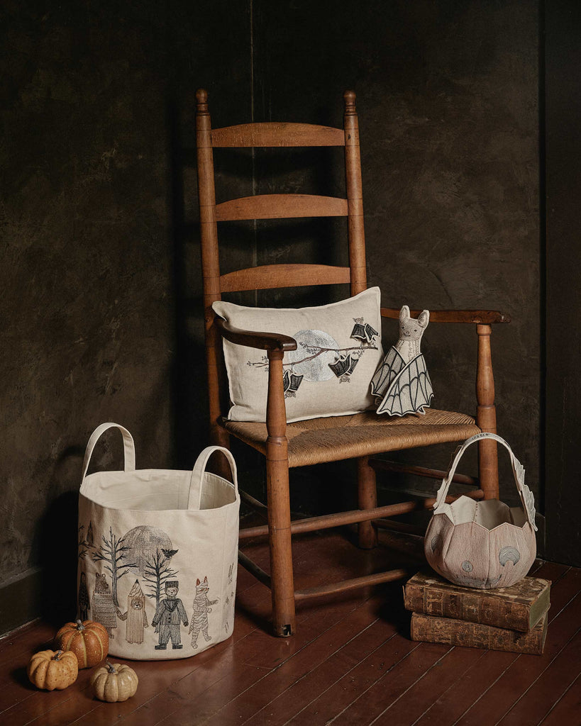 A rustic wooden rocking chair holds the Coral & Tusk Bat Branch Pillow and a doll in a bat costume, creating a charming Halloween display. A nearby decorated basket with animal illustrations completes the cozy scene.