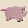 The Holztiger Piglet is a hand-carved pink wooden piglet with a curly tail, black hooves, and a small smile, shown on a beige background. Handcrafted in Europe and ships in one week.