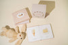 On a light background, a stack of children's books and a beige plush rabbit create a charming scene. One book is open with "Ice Cream" and "Cake" labeled images. Nearby, the cube-shaped box titled "Our Little Library Vol. 2" perfectly complements this delightful board book set. Shadows gently fall across the surface.