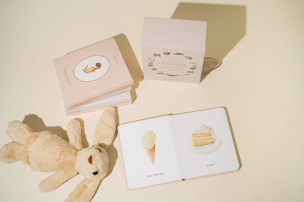 On a light background, a stack of children's books and a beige plush rabbit create a charming scene. One book is open with "Ice Cream" and "Cake" labeled images. Nearby, the cube-shaped box titled "Our Little Library Vol. 2" perfectly complements this delightful board book set. Shadows gently fall across the surface.