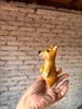 A hand holds a small, handmade wool felt Shiba-Inu Dog Finger Puppet against a background of white-painted brick wall.