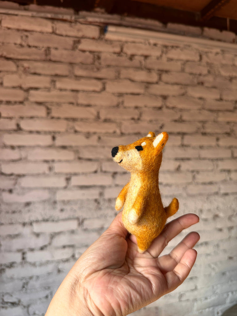A hand holds a small, handmade wool felt Shiba-Inu Dog Finger Puppet against a background of white-painted brick wall.