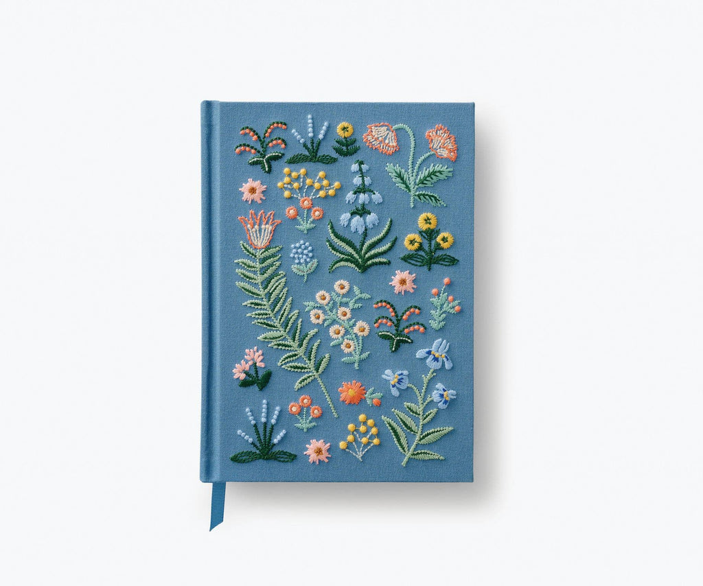 The Menagerie Garden Embroidered Journal features a light blue hardcover with colorful floral and leaf embroidery on the front, plus a matching blue ribbon bookmark.