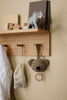 The Ferm Living Place Rack - Small, crafted from oak, organizes books, a white net bag, an elephant figurine, a hanging plush bear toy with ring, and a white shirt on pegs against a beige wall for stylish home storage.