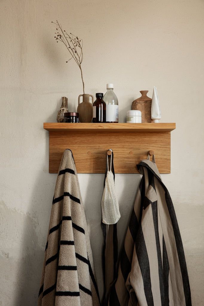 The Ferm Living Place Rack - Small, styled with bottles, jars, and vases on top and three striped towels plus a mesh bag hanging below, is perfect for chic home organization against a beige wall.