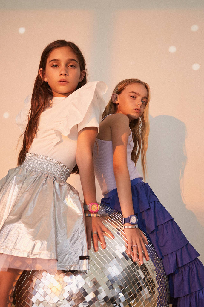 Two girls in stylish outfits lean against a large disco ball, each wearing a Mini Kyomo Watch - Disco. These kids watches add a playful touch as they pose confidently under soft lighting.
