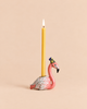A yellow beeswax birthday candle sits in the Flamingo Cake Topper - Final Sale (ships in one week), a ceramic flamingo with a blue and green party hat, against a solid light beige background.