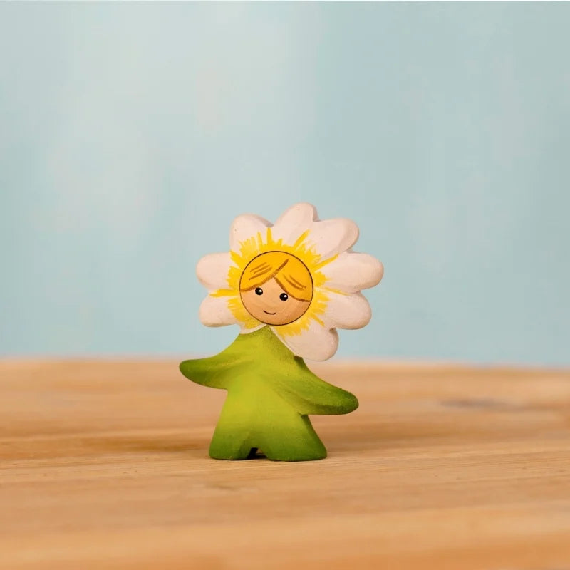 The Bumbu Handmade Wooden Flower Child Daisy, featuring a smiling face amid white petals and a green leafy body, stands on light wood with a blue backdrop—ideal for Montessori learning and fostering a connection to nature.