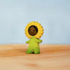 The Bumbu Handmade Wooden Flower Child Sunflower features a small, smiling figurine with yellow petals and green attire, standing on a light wooden surface against a pale blue background. Ships in one week.