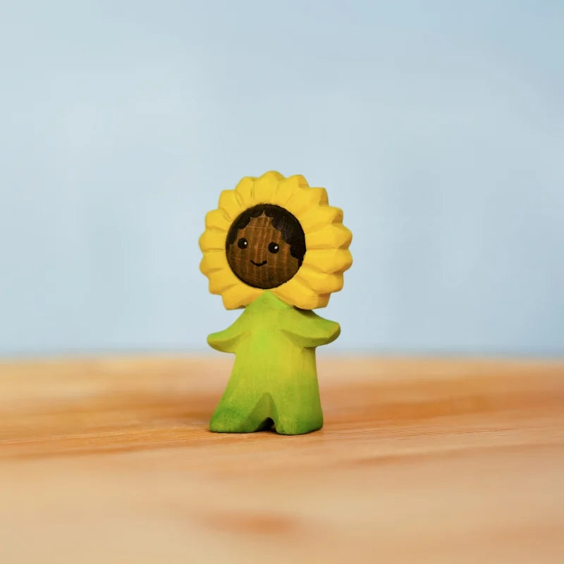 The Bumbu Handmade Wooden Flower Child Sunflower features a small, smiling figurine with yellow petals and green attire, standing on a light wooden surface against a pale blue background. Ships in one week.