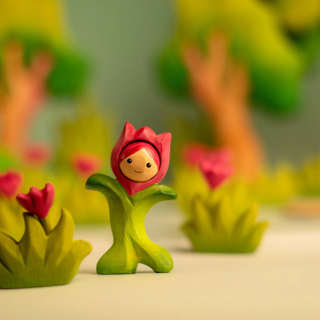 The Bumbu Handmade Wooden Flower Child Tulip, featuring a smiling face and red petal hat, stands among green and pink plants—inviting open-ended play—against a blurred background of trees and flowers. Ships in one week.