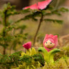 The Bumbu Handmade Wooden Flower Child Tulip, featuring a smiling face, sits on green moss among pine branches. A red flower bud and a blurred butterfly enhance this inviting scene for open-ended play. Ships in one week.