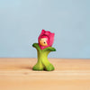 The Bumbu Handmade Wooden Flower Child Tulip, a smiling wooden toy with green leaves and a pink blossom, invites open-ended play as it sits on a light surface against a blue background. Ships in one week.