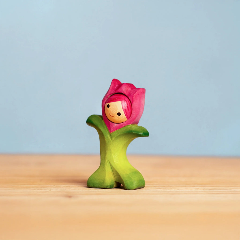 The Bumbu Handmade Wooden Flower Child Tulip, a smiling wooden toy with green leaves and a pink blossom, invites open-ended play as it sits on a light surface against a blue background. Ships in one week.